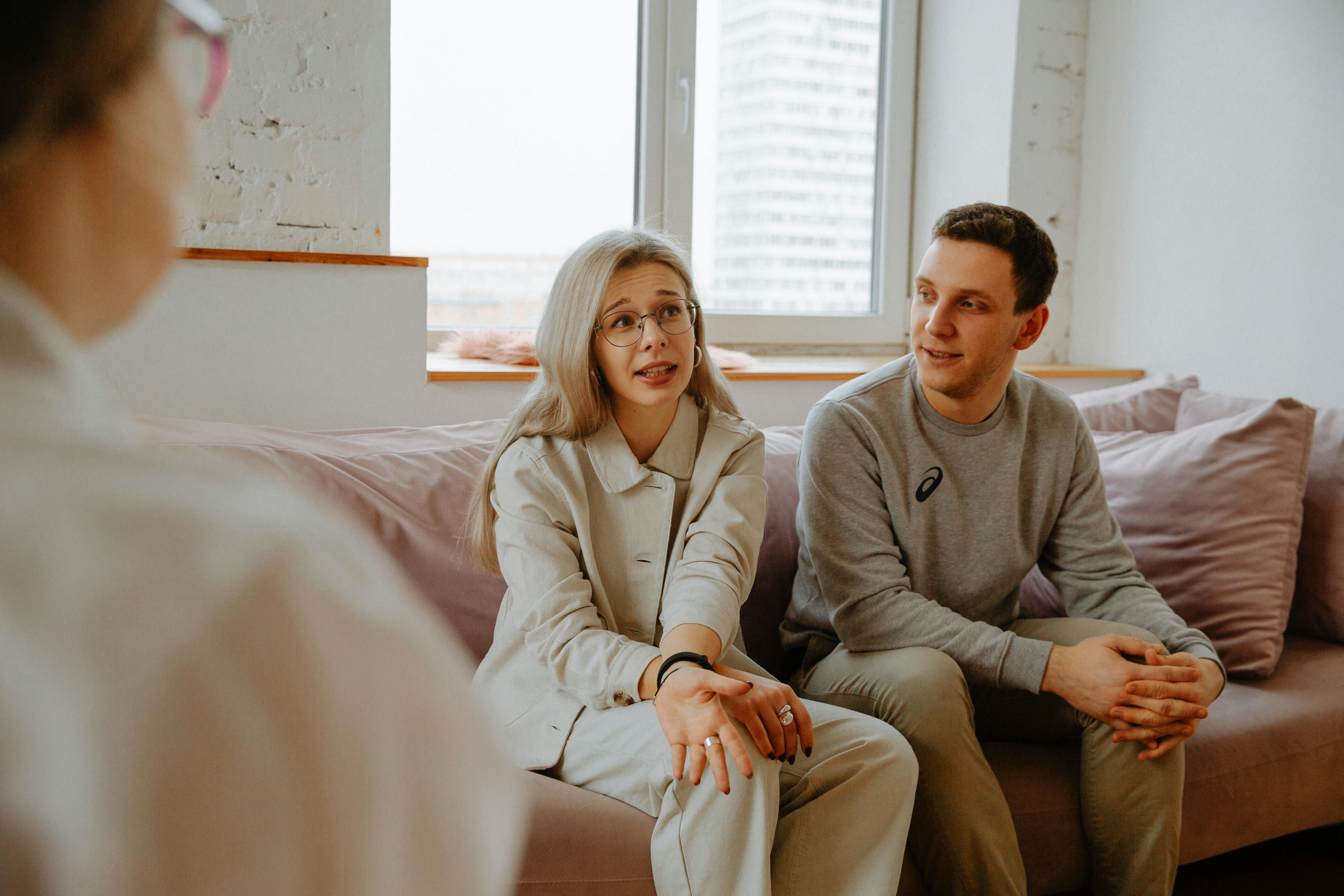 Caucasian couple on couch talking to a caucasian person.