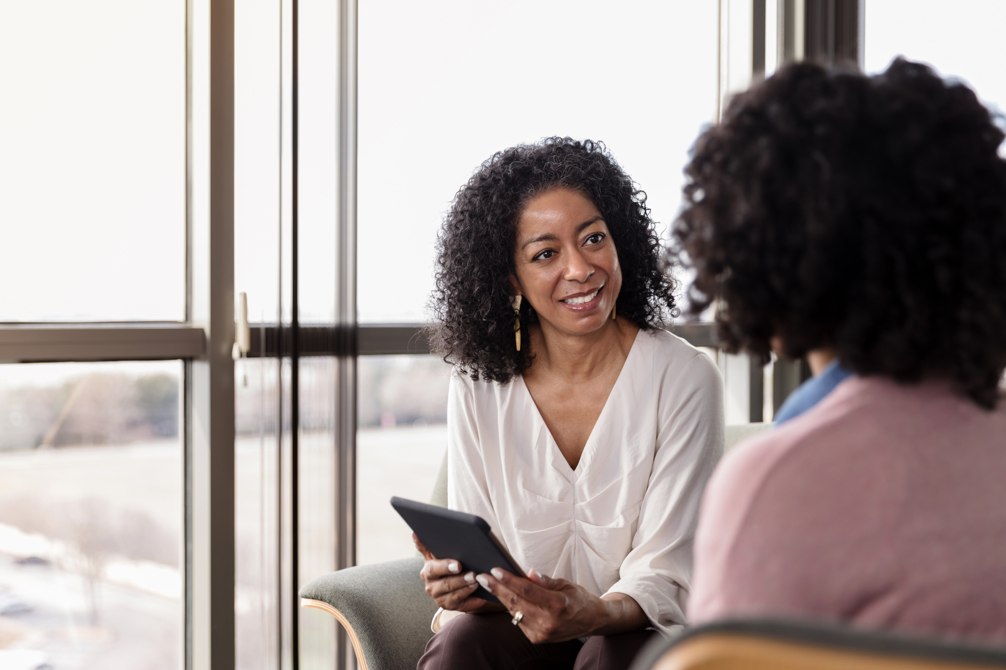 Two African American women facing each other, one has a smile and is holding a tablet. The other has her back to us. 