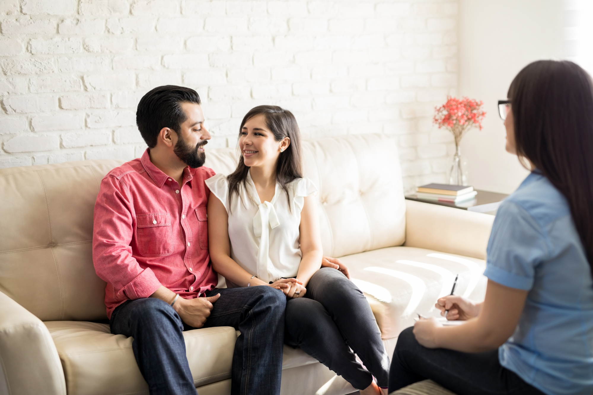 Couple sitting on a couch smiling at one another with a person sitting across from them holding a pen and pad of paper.