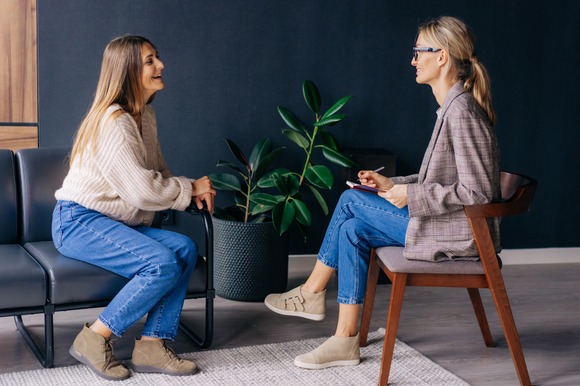 Two Caucasian women sitting across from one another. One is holding a pen and notepad, the other is smiling and talking.