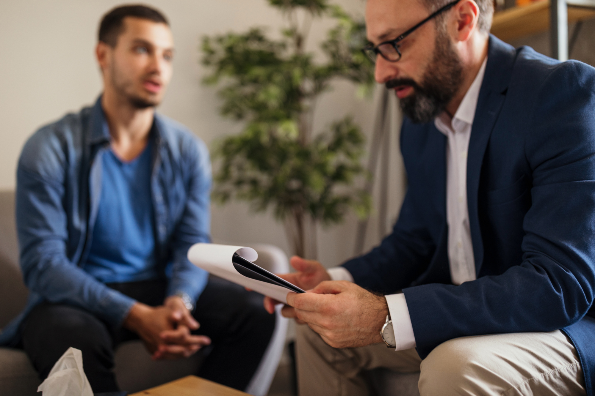 Man in background talking with man in front looking at notepad