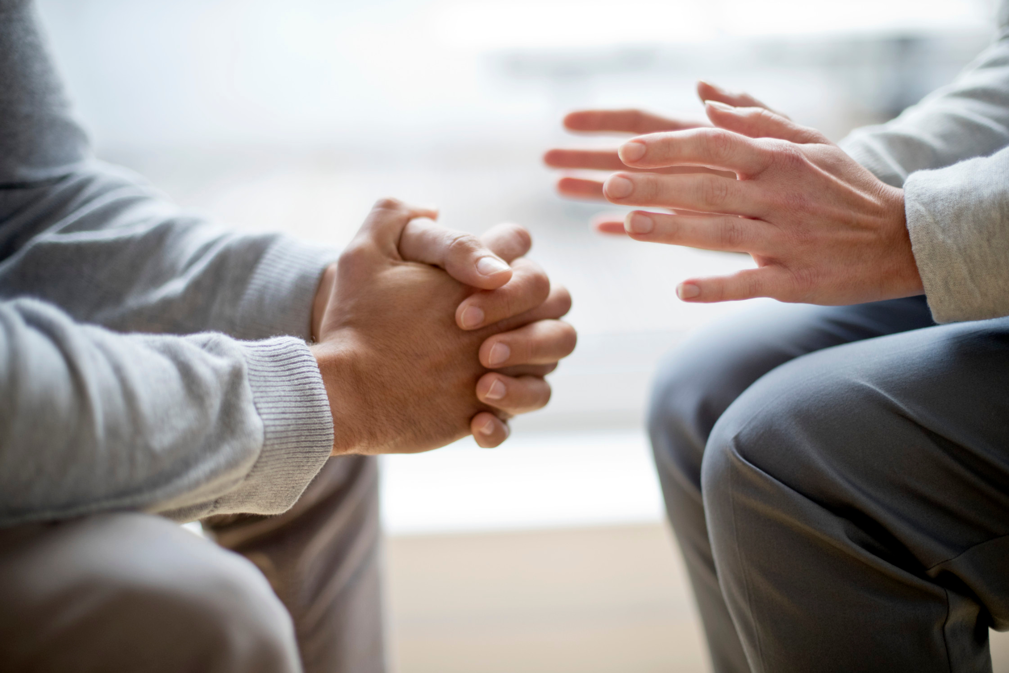 Close up of two people sitting facing one another. One has their hands clasped together, the other has their hands in motion as if talking.