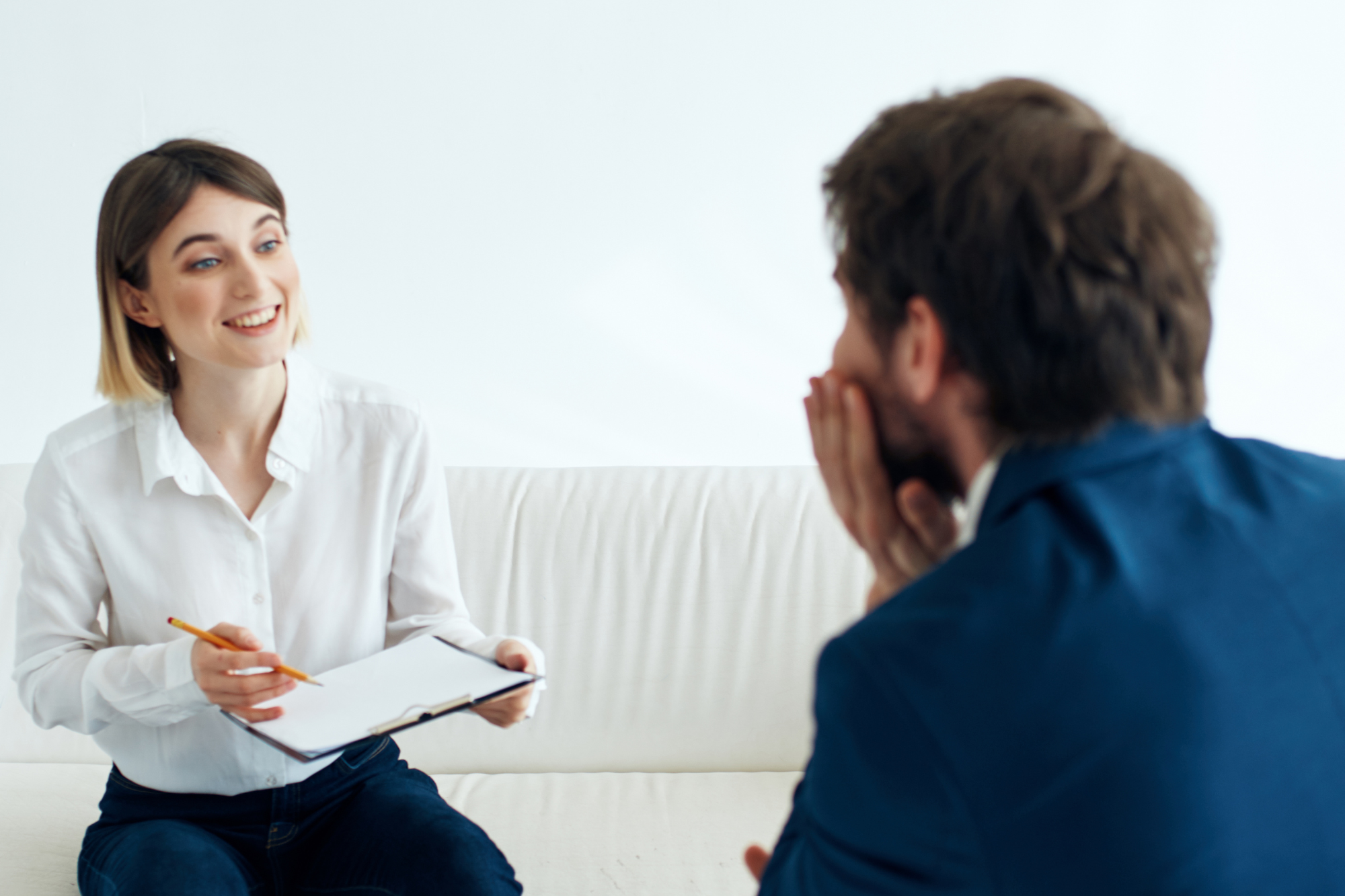 Caucasian woman sitting on a couch with a clipboard and pencil and smiling at a Caucasian man in suit sitting across from her.